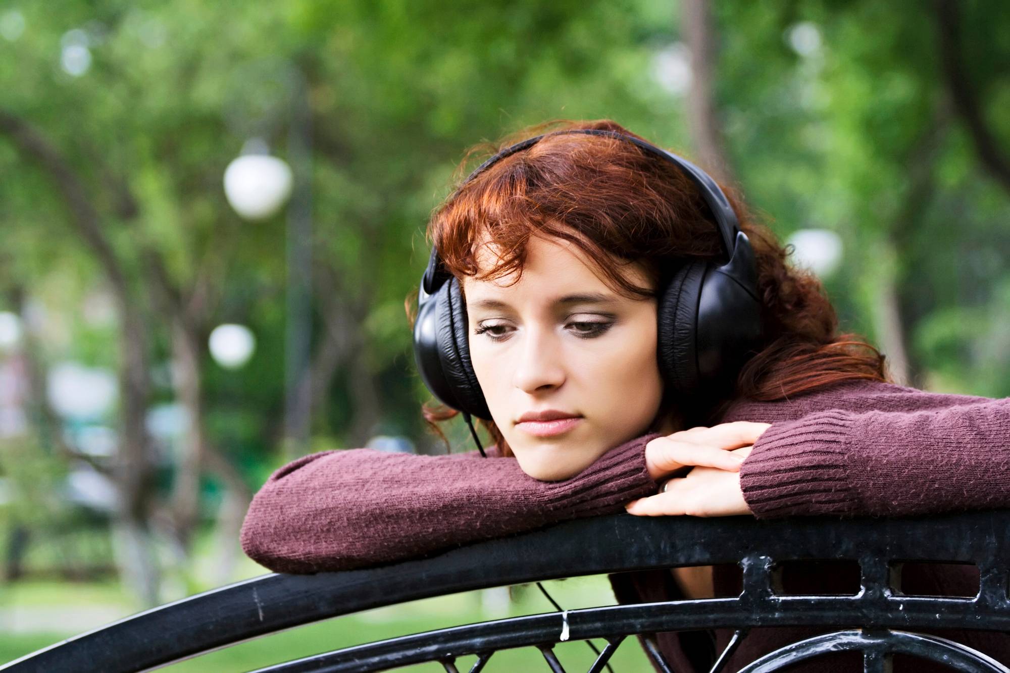 Woman wearing over-the-ear headphones listening to music with her head resting on the back section of a park bench